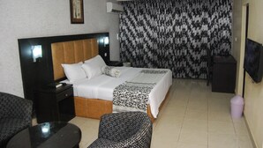 A hotel room with a bed, a nightstand, a chair, a small table, a television, and a curtain with a black and white pattern. - DAWN CARRINGTON HOTELS AND SUITES (Lagos)