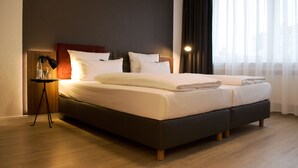 Uma cama bem arrumada com roupa de cama branca, uma mesa de cabeceira com abajur e uma janela com cortinas finas.
