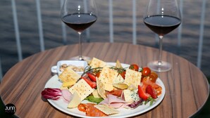 Una mesa de madera con un plato de quesos, carnes y galletas, acompañado por dos copas de vino tinto, sobre un fondo desenfocado.