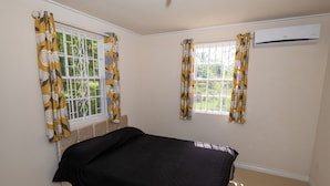A bedroom with a bed, two windows with curtains, an air conditioning unit, and a view of greenery outside. - THE Cottage, Saint Davids, Christ Church, Barbados (Oistins)