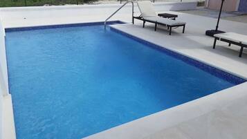 Piscina retangular com borda azulejada, escada de metal e duas cadeiras de descanso com uma pequena mesa.