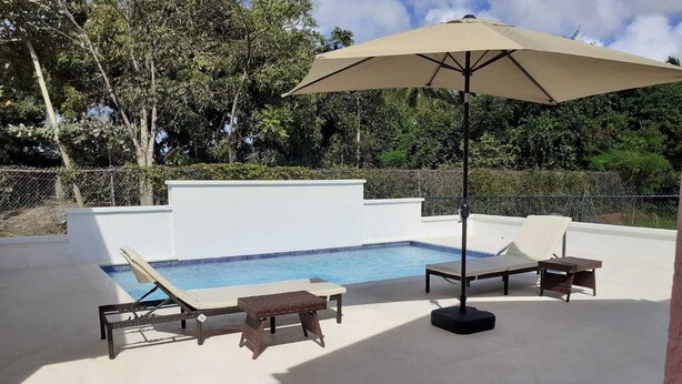 An outdoor swimming pool with lounge chairs and a table, surrounded by a white wall and greenery. - THE Cottage, Saint Davids, Christ Church, Barbados (Oistins)