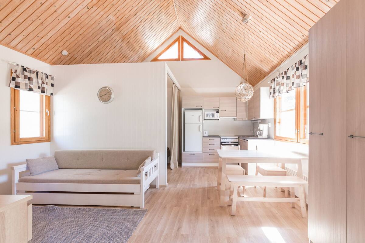 A spacious room with a high wooden ceiling, a sofa, a dining table, and a kitchen area.