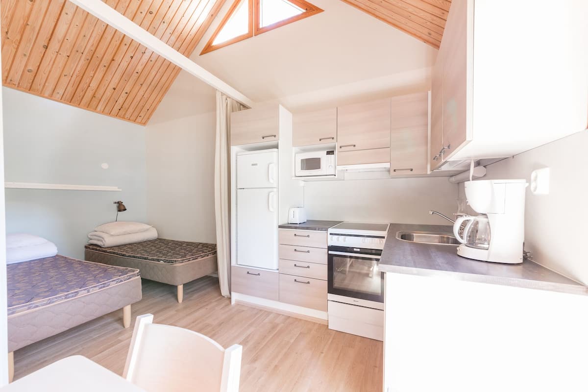 A compact kitchenette with a microwave, toaster, coffee maker, and stove. There is a bed with a patterned mattress and pillows, and a small seating area with a chair.