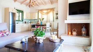A cozy living room with a fireplace, a dining area, and a kitchen with wooden beams. - Ktima Aniforeli Luxury Villas (Lefkada)