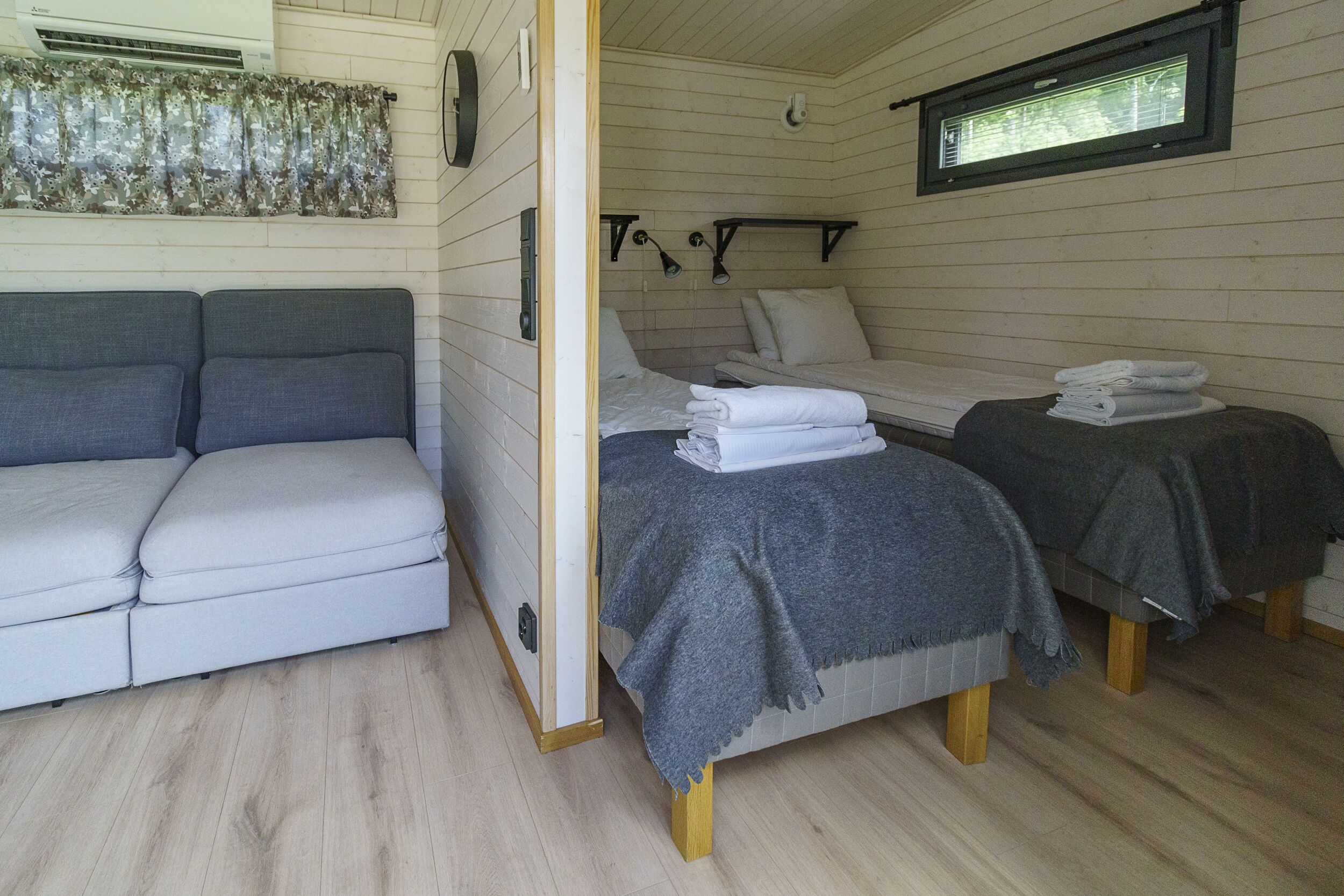 A room with a sofa, two beds with folded towels, a window, and a wall-mounted air conditioner.