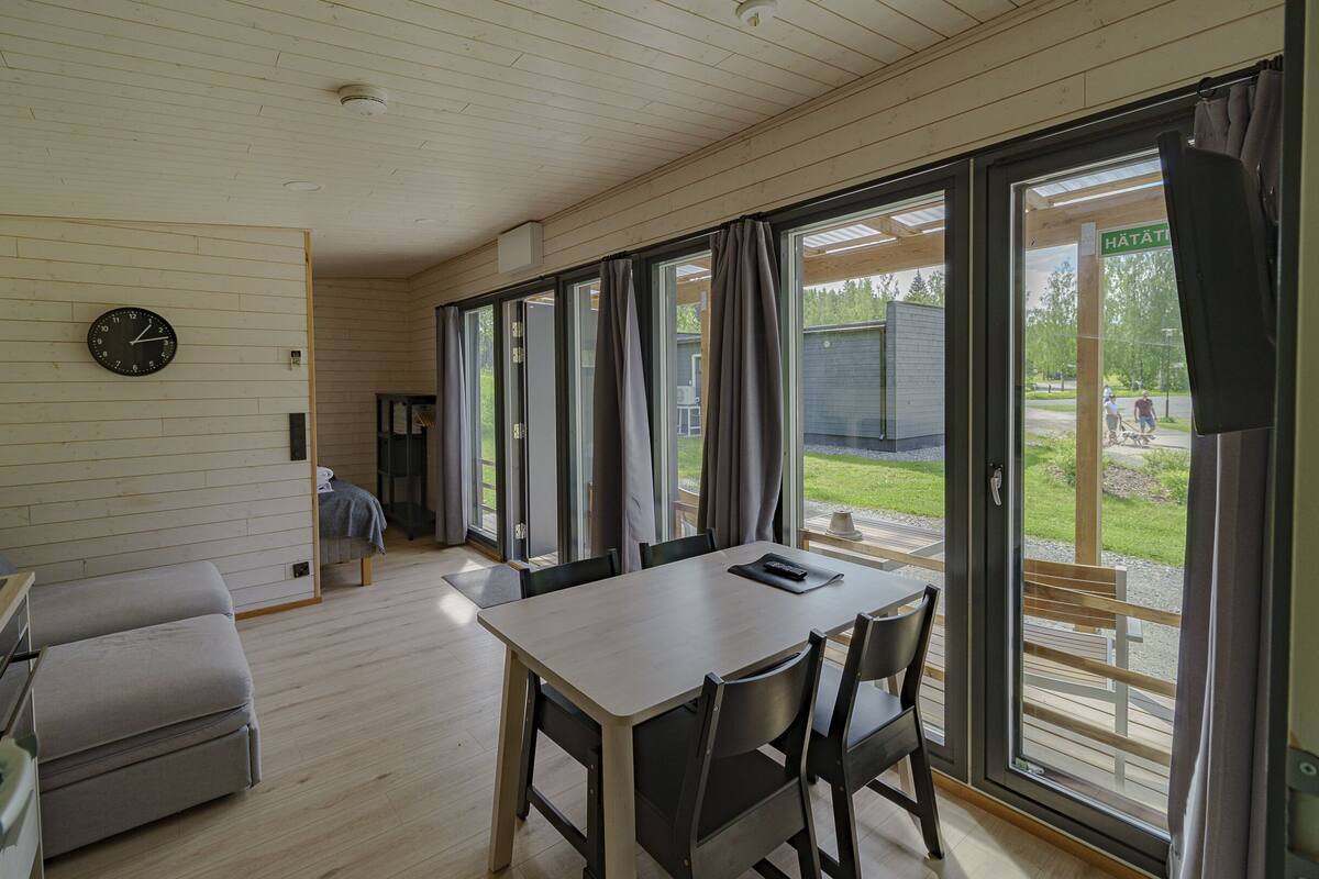 A room with a wooden floor, a table with chairs, a sofa, a clock on the wall, and large sliding glass doors leading to an outdoor area.