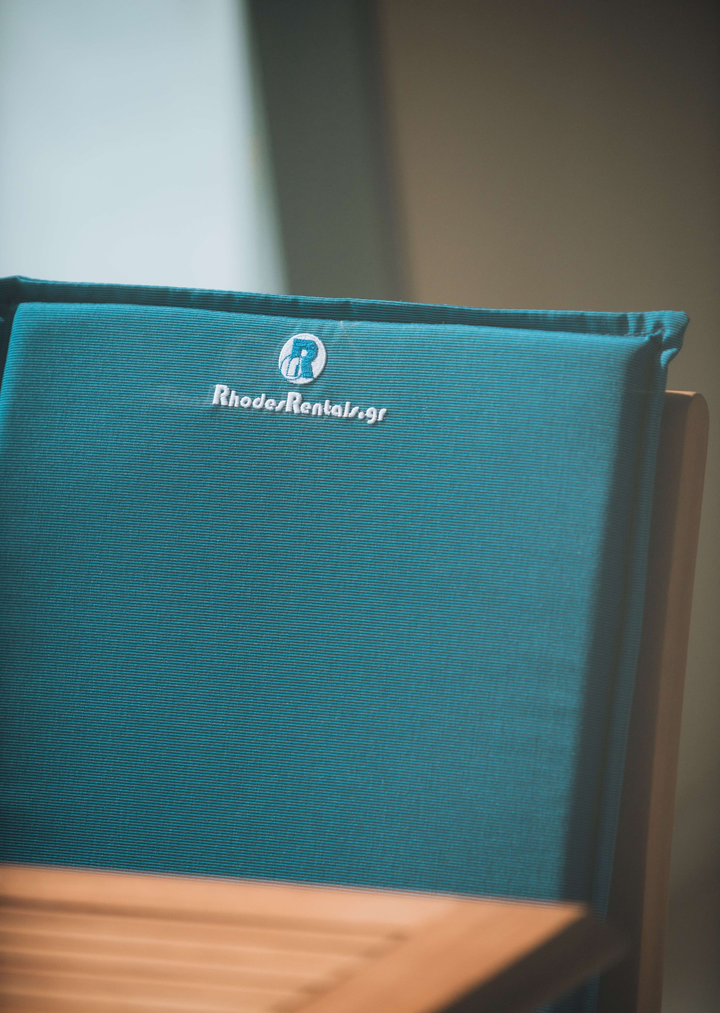 Una silla de tela color aguamarina con un logotipo y el texto "RhodesRentals.gr".