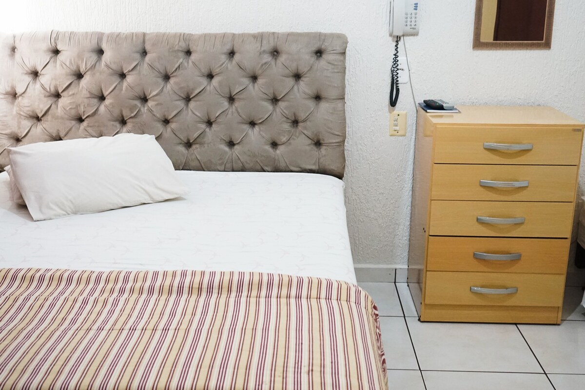 A bed with a tufted headboard, a bedside table with drawers, a telephone, and a striped area rug.