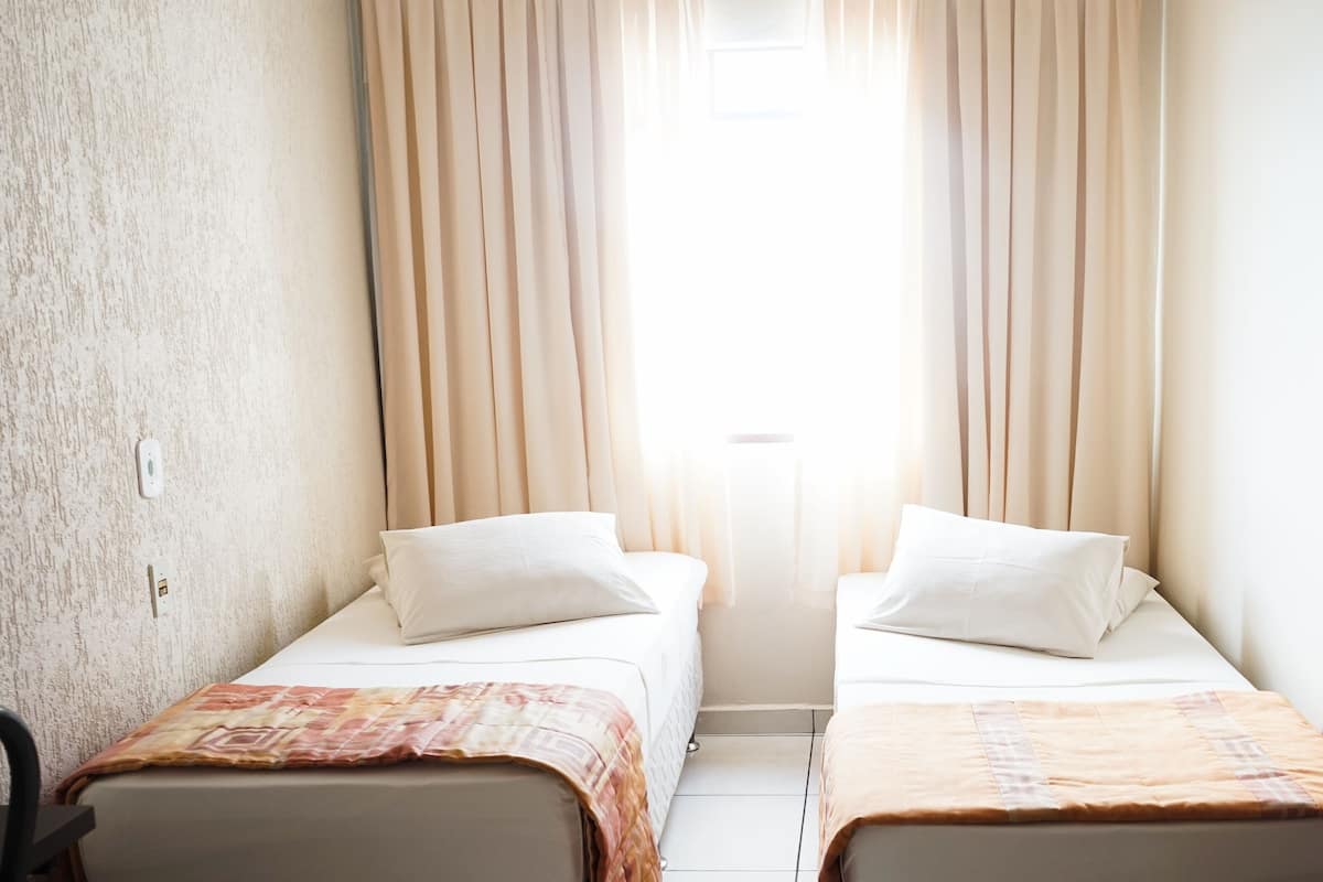 A small, bright room with two beds, beige curtains, and a window.