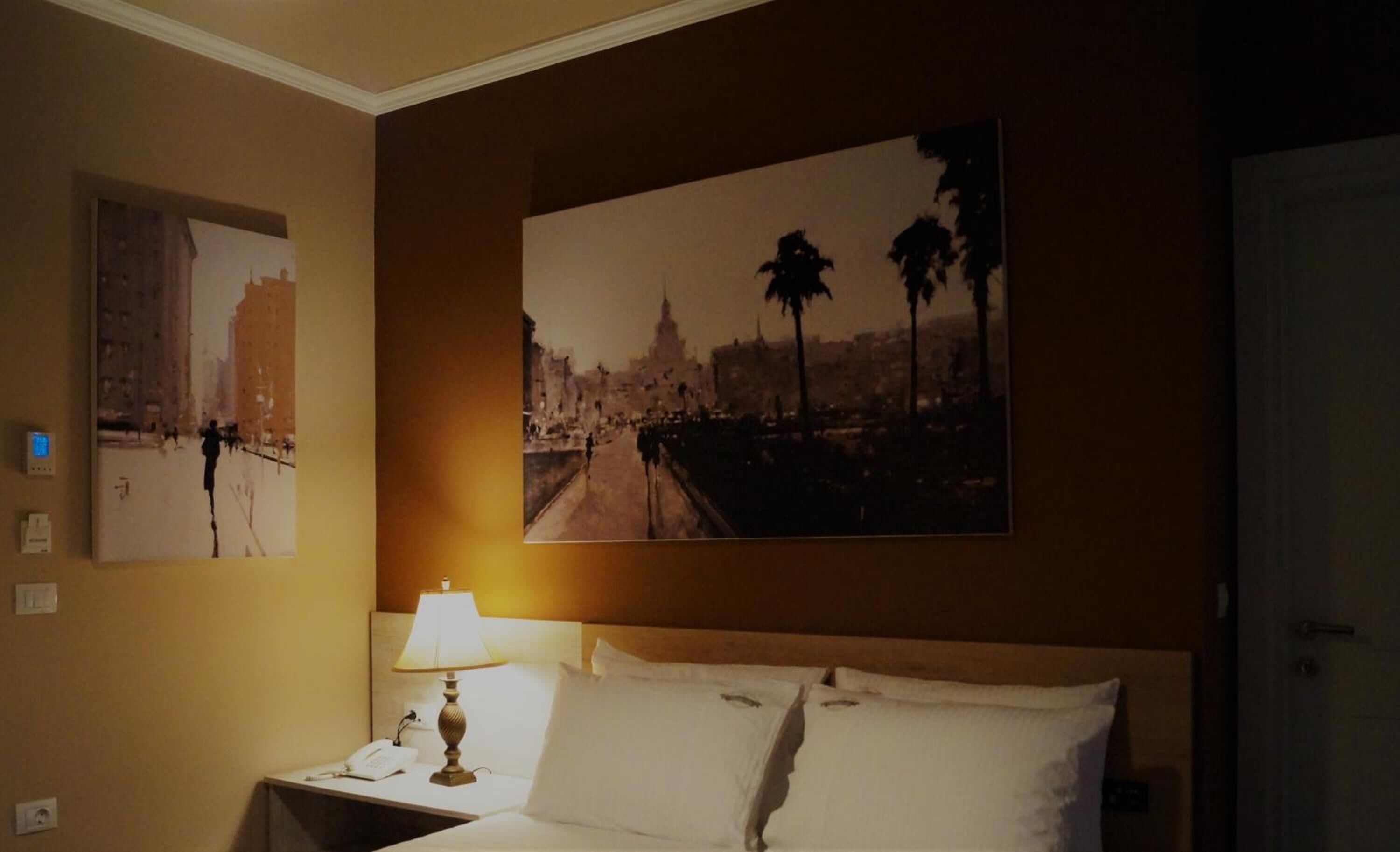 Habitación de hotel con cama, mesita de noche con lámpara, teléfono y dos cuadros urbanos colgados en la pared.