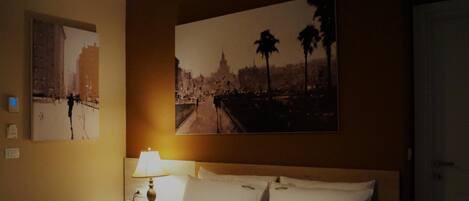 Habitación de hotel con cama, mesita de noche con lámpara, teléfono y dos cuadros urbanos colgados en la pared.