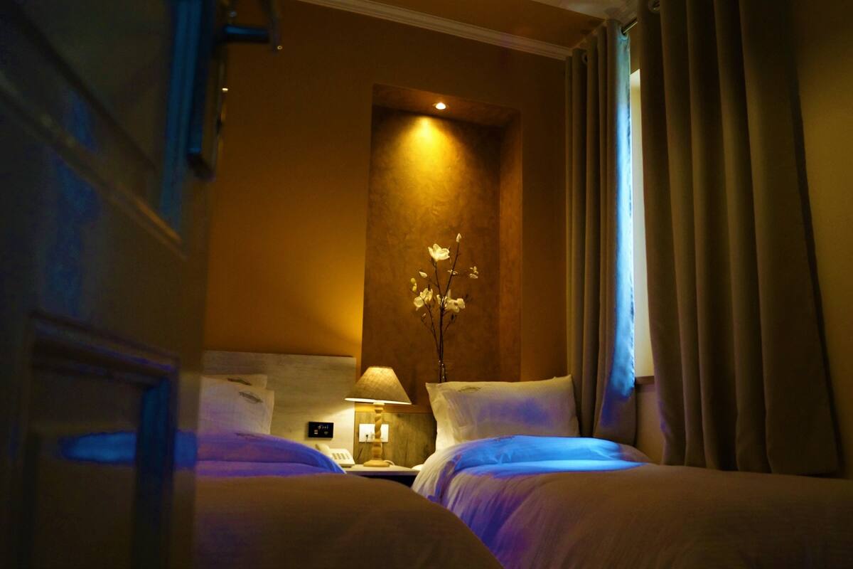 A hotel room with two beds, a nightstand with a lamp, a vase with flowers, and a window with curtains.