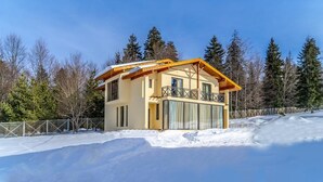 Una cabaña de dos pisos con balcón, rodeada de nieve y árboles.