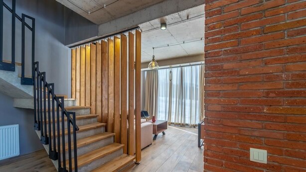 Una escalera de madera con barandillas negras que conduce a una habitación con pared de ladrillo y una ventana con cortinas.