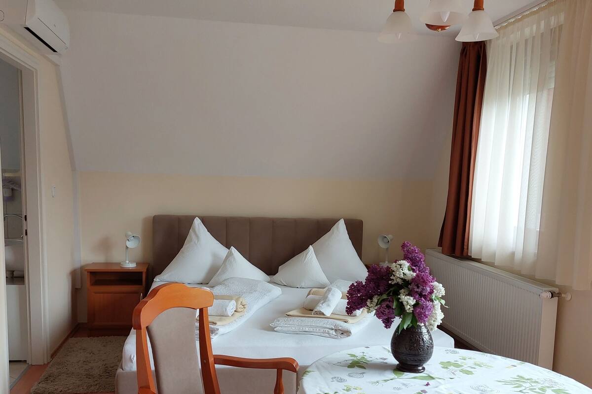 A hotel room with a bed, a wooden chair, a table with a floral tablecloth, and a vase with flowers.