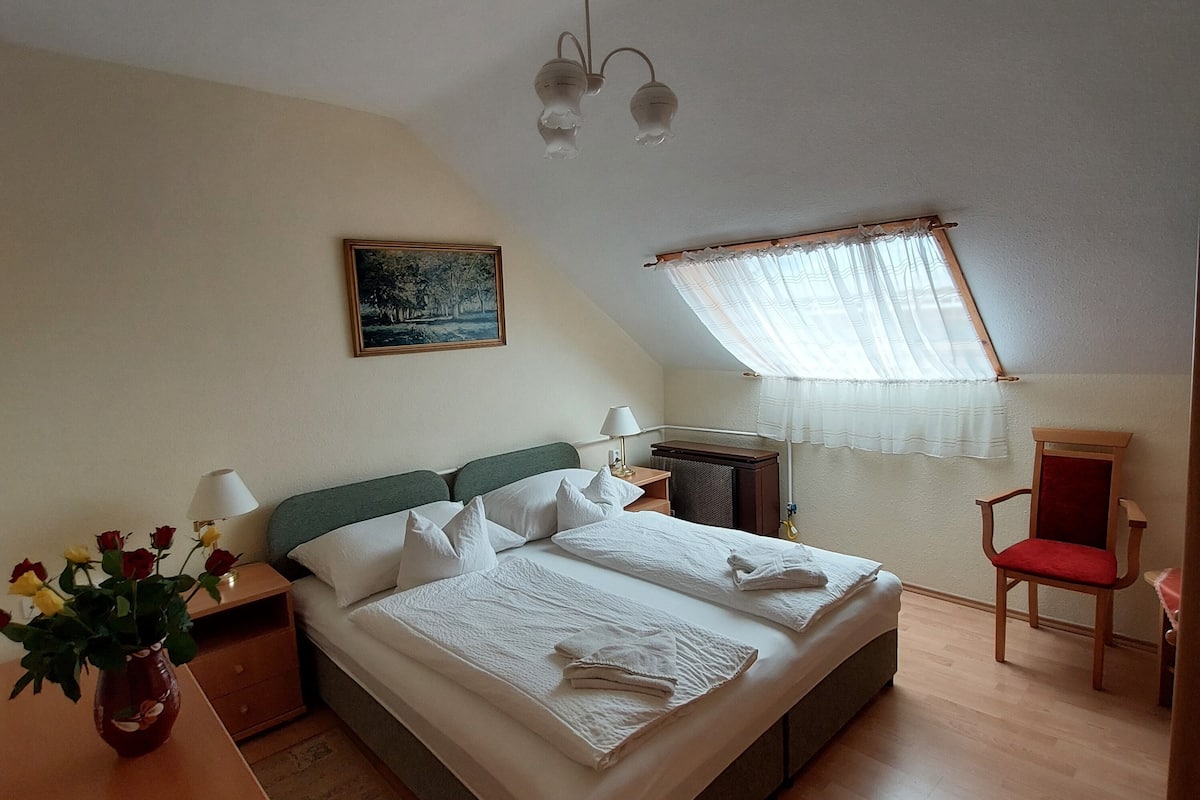 A neatly arranged bedroom with a bed, bedside tables, a chair, a window with curtains, and a painting on the wall.