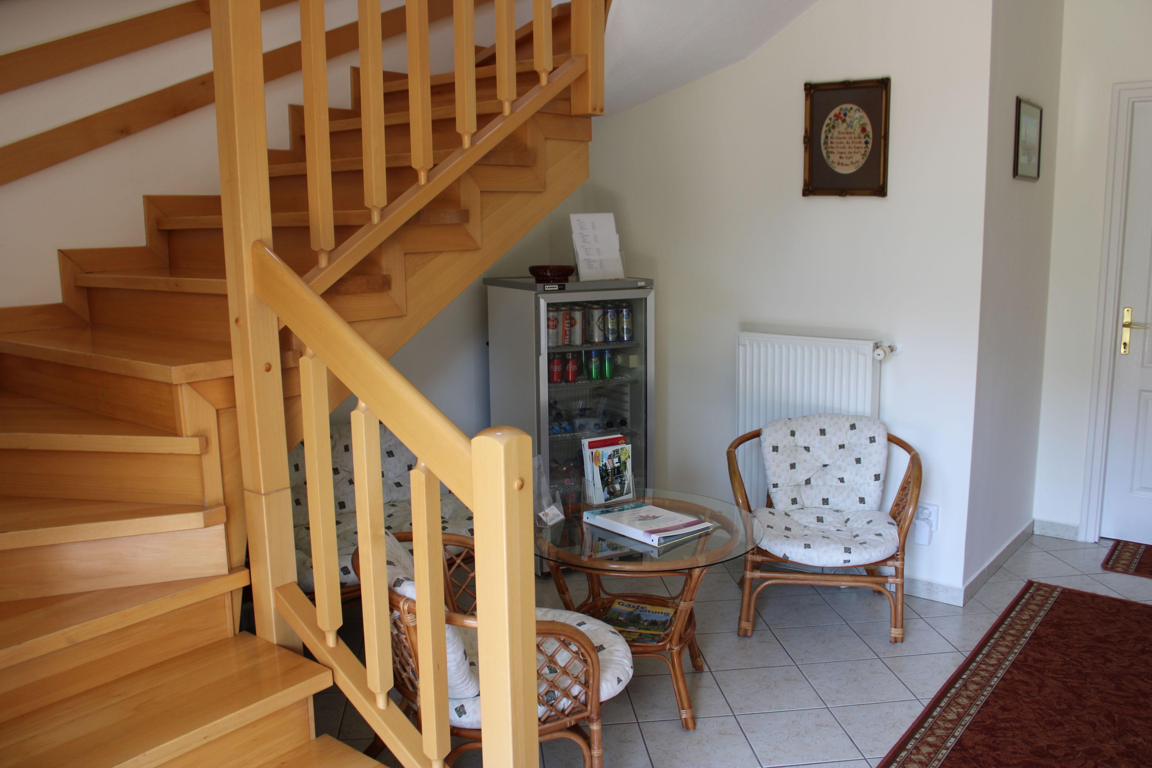 A wooden staircase leading to a landing with a radiator, a wicker chair, a small table with a magazine, and a refrigerator.