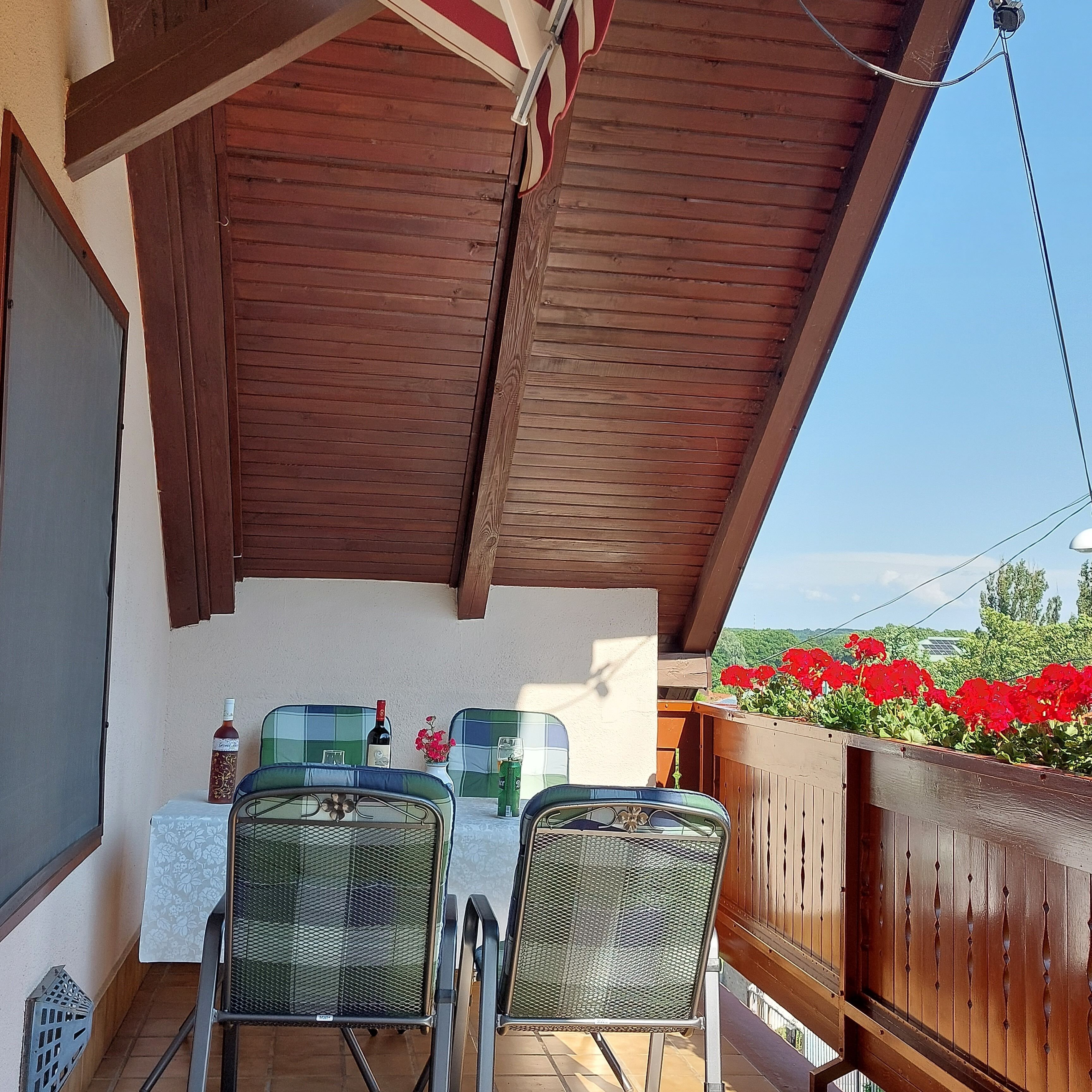 A covered outdoor seating area with two metal chairs, a table, and a bottle of wine.