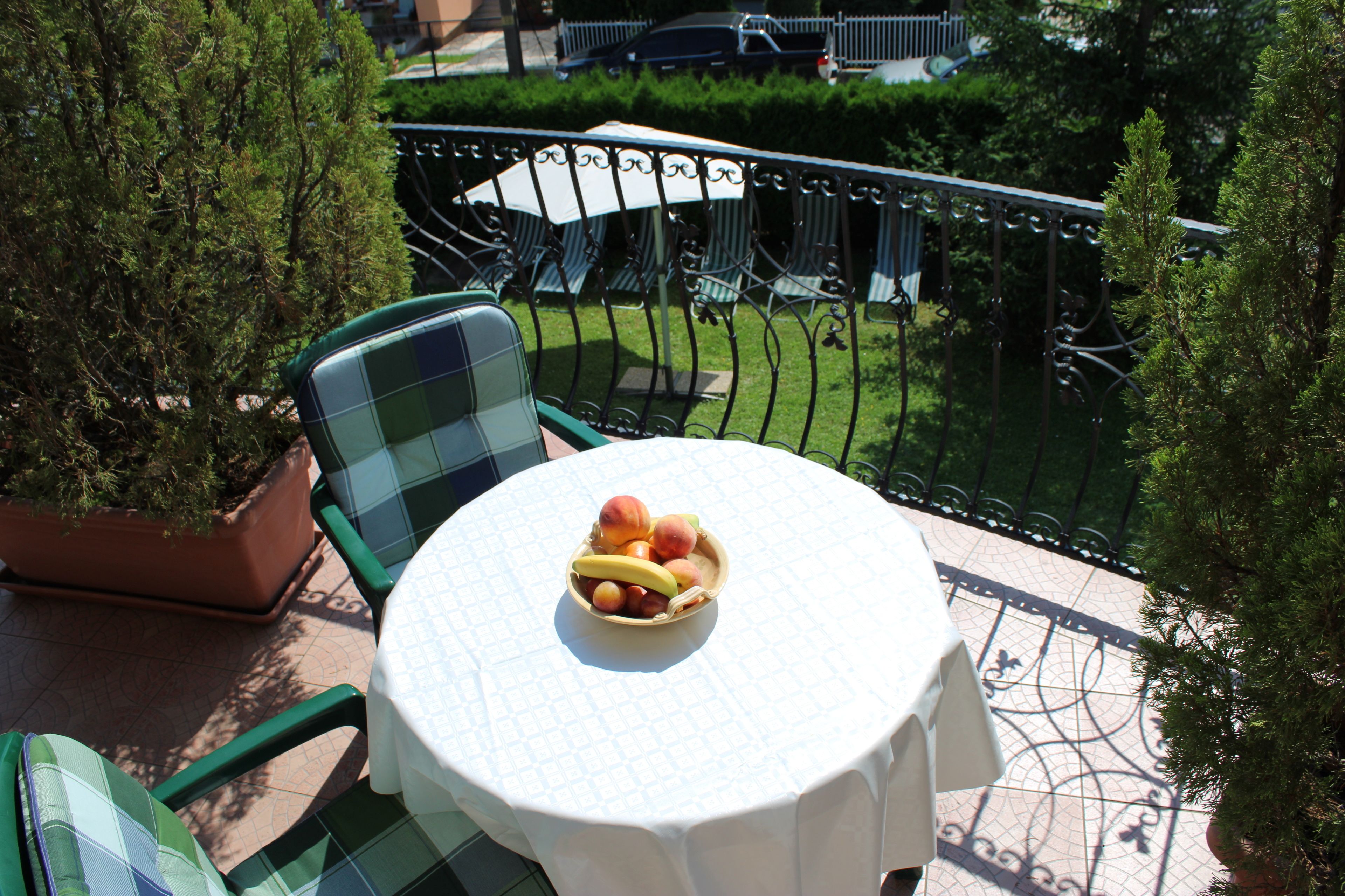 A round table with a white tablecloth, a bowl of fruit, and green chairs on a balcony with a view of a garden and a fence.