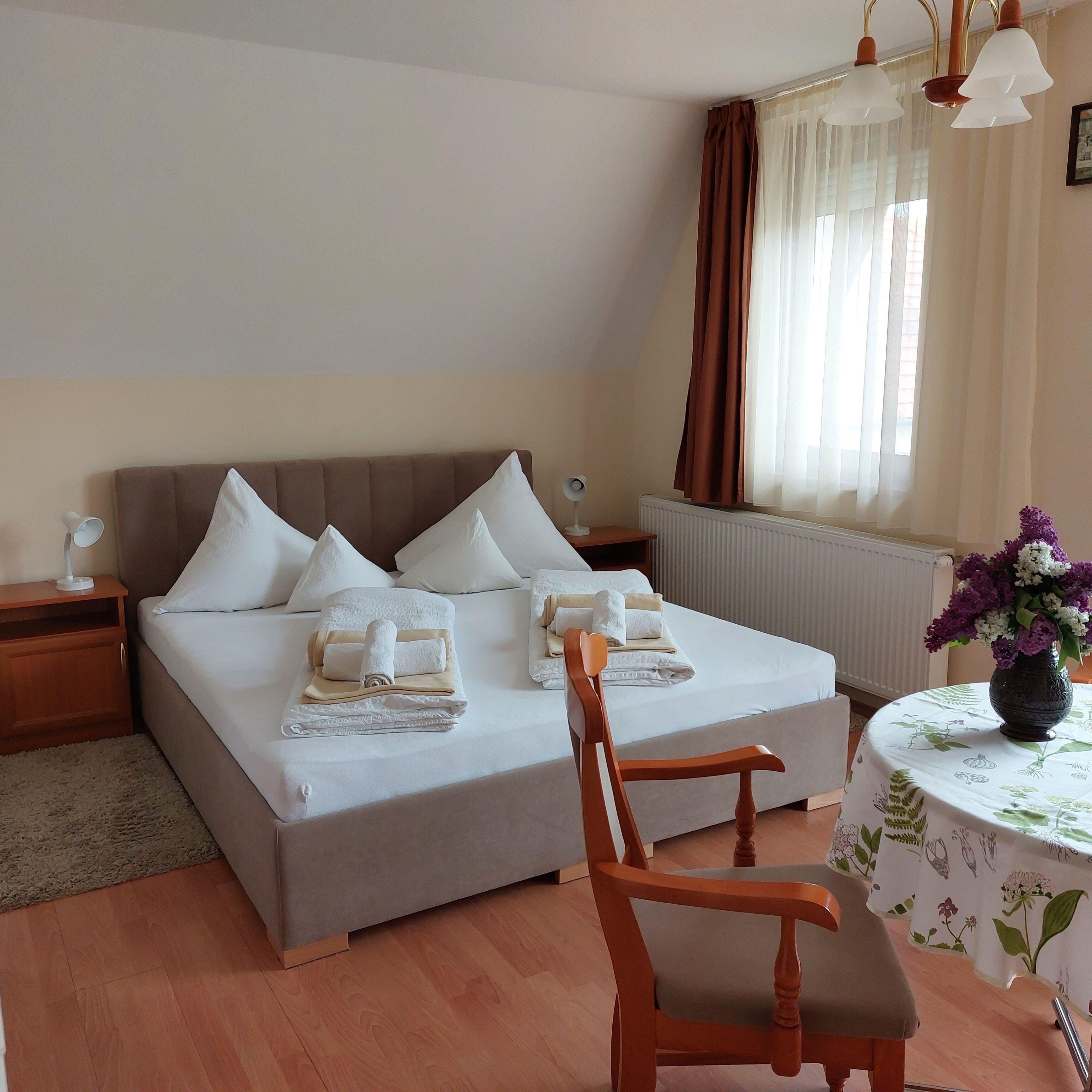 A hotel room with a double bed, a wooden chair, a small table with a vase of flowers, and a nightstand with a lamp.