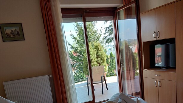 A hotel room with a bed, a TV, a window with curtains, and a view of the outside. - Erika Apartmanházak (Hévíz)