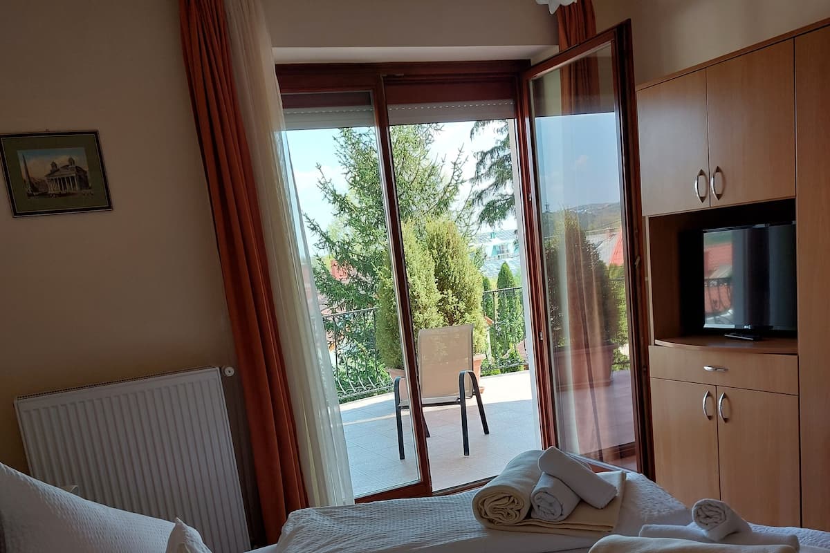 A hotel room with a bed, a TV, a window with curtains, and a view of the outside.