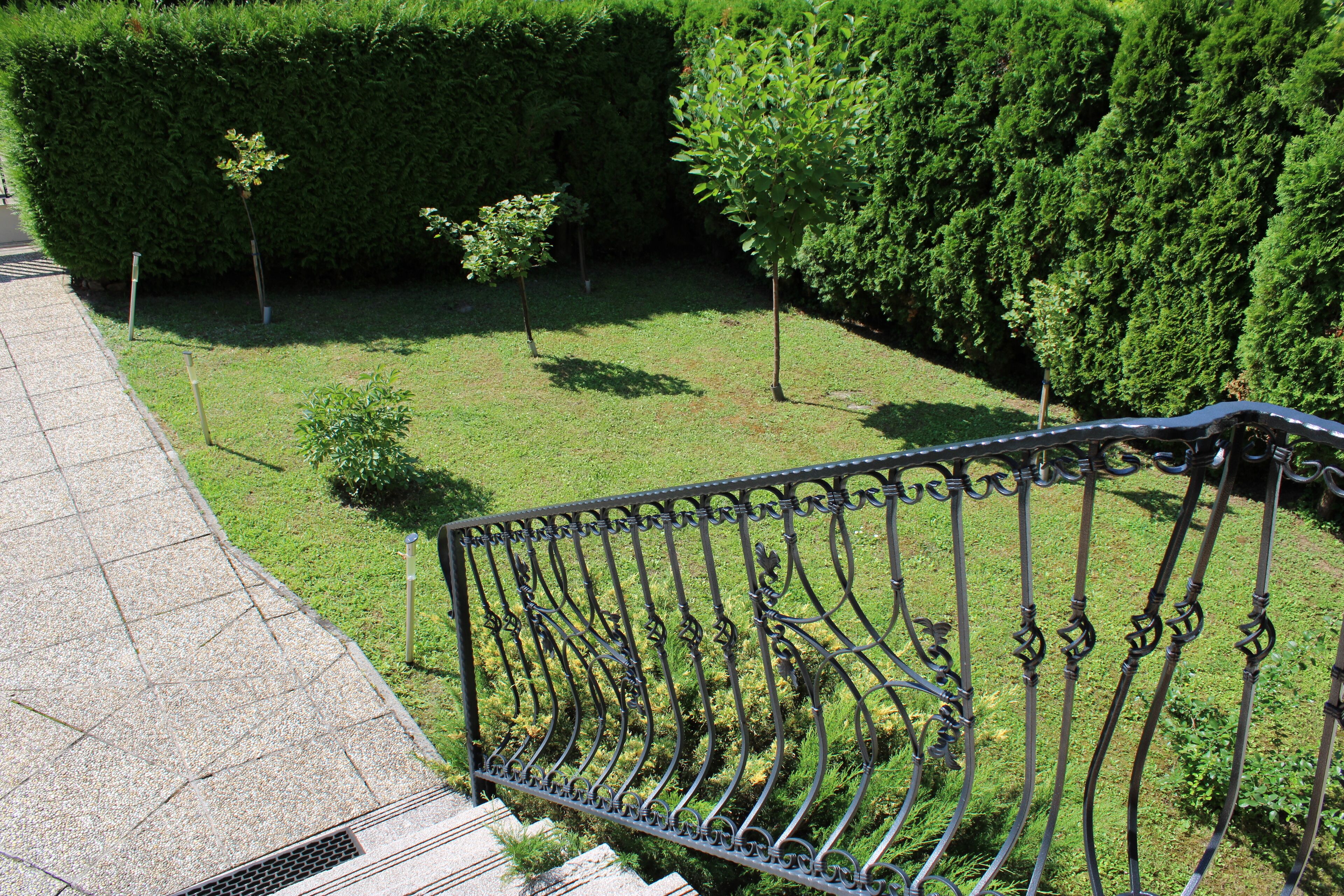 A garden with a metal fence, a paved walkway, and various green plants.