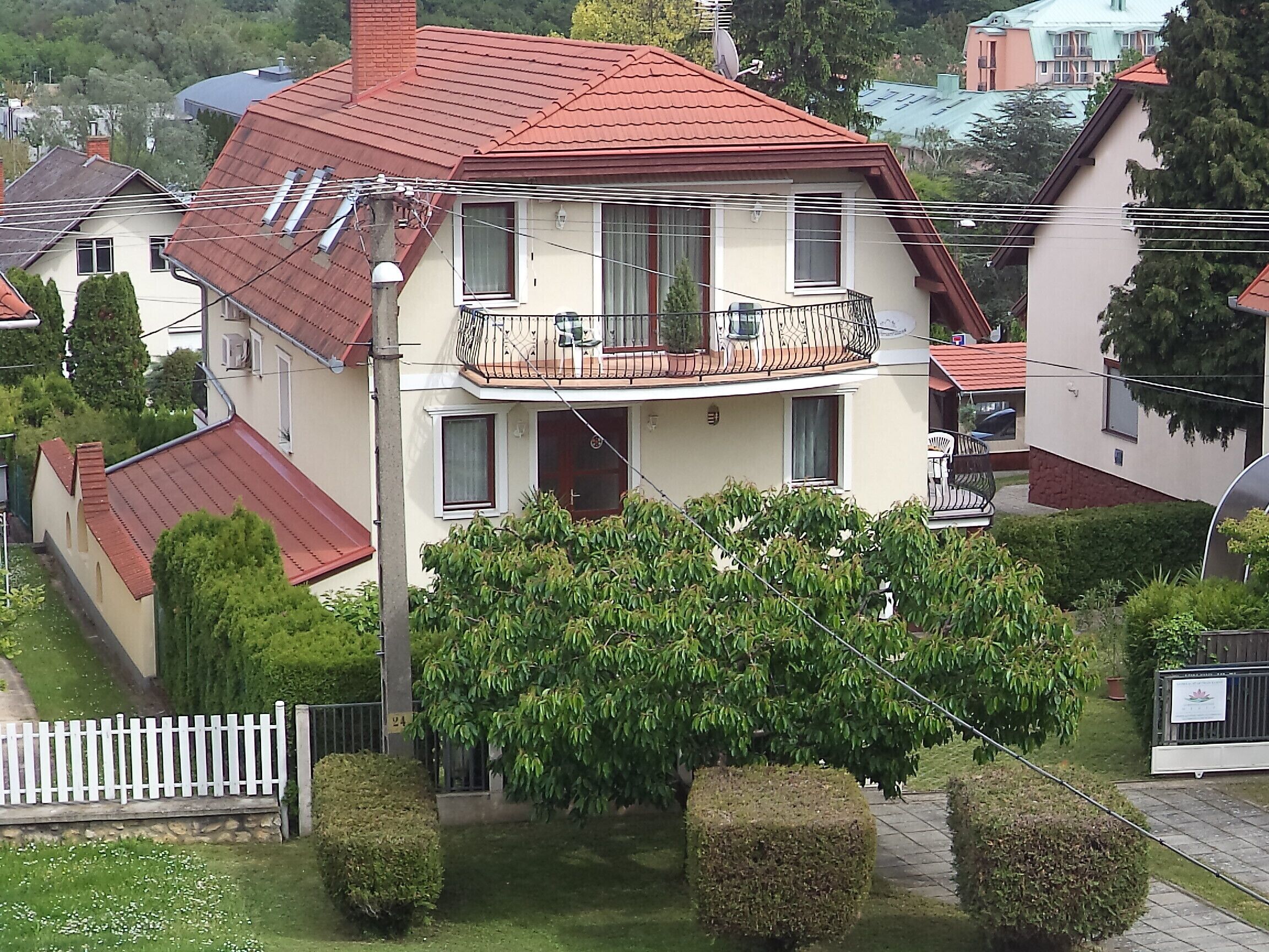 Una casa de dos pisos con techo rojo, un balcón con barandilla y un jardín bien cuidado con setos.