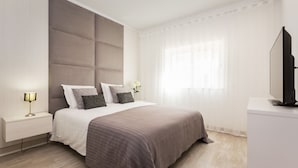 A modern bedroom with a large bed, a bedside table with a lamp, a flat-screen TV on a stand, and a large window with sheer curtains. - D Loft Downtown (Albufeira)