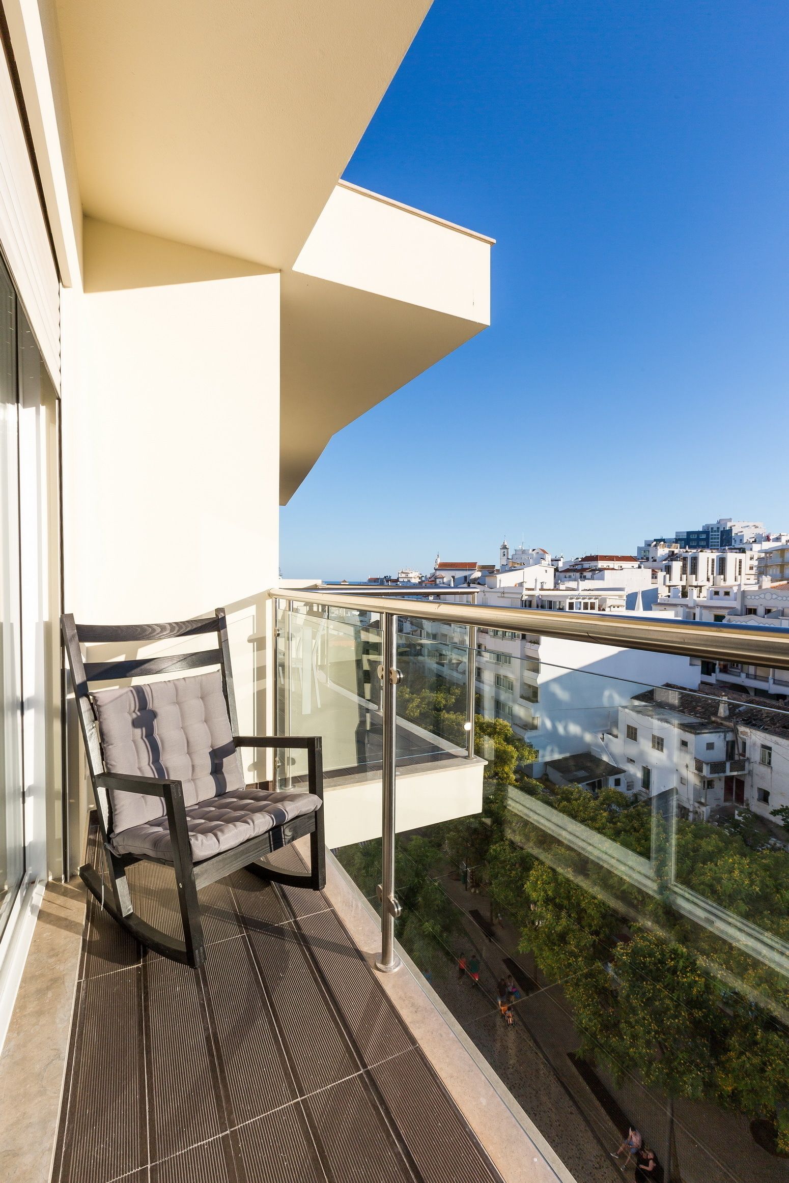A balcony with a glass railing, a cushioned chair, and a view of a cityscape.