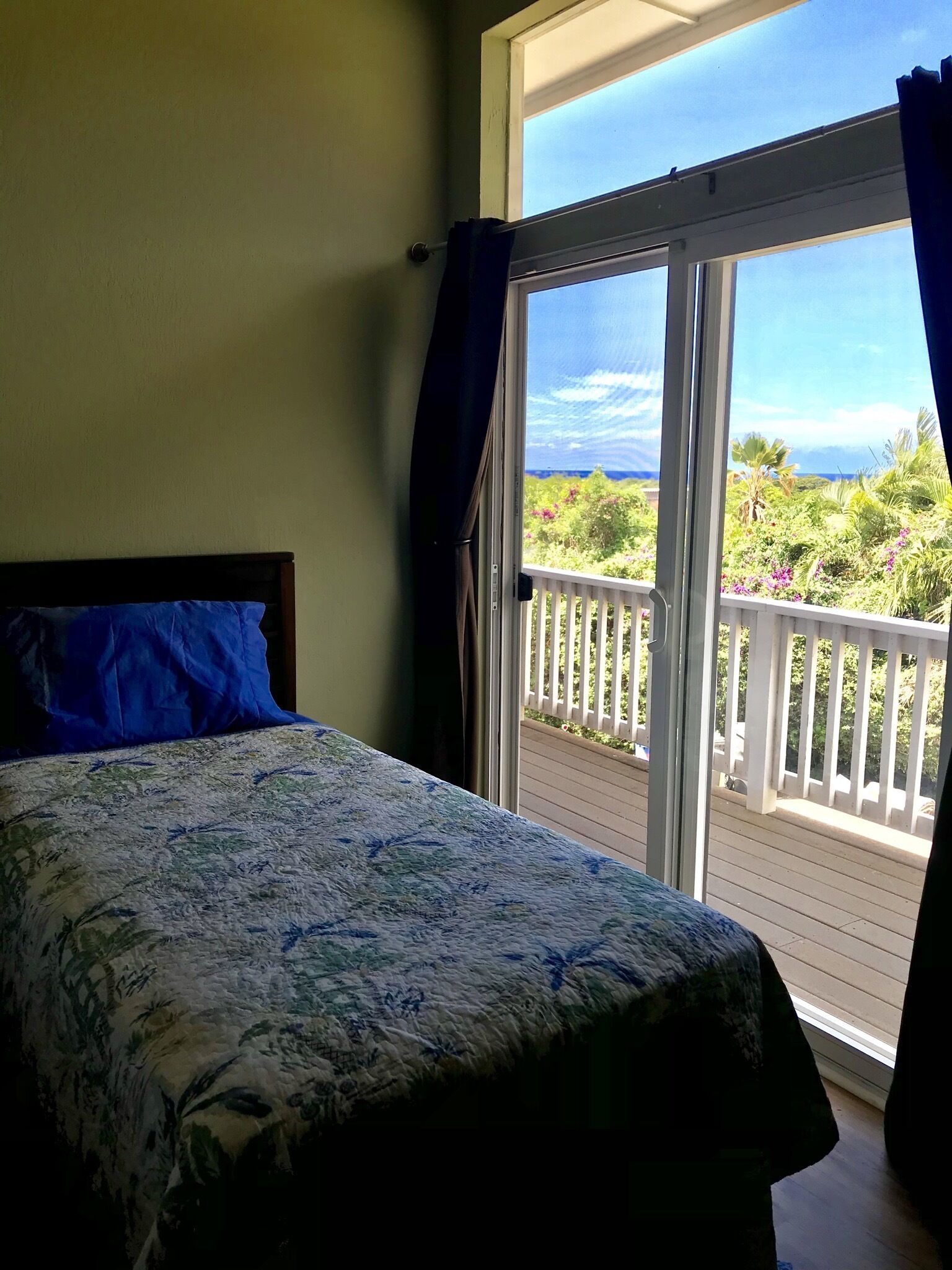 A bedroom with a bed, a window with curtains, and a view of a balcony with plants and a railing.