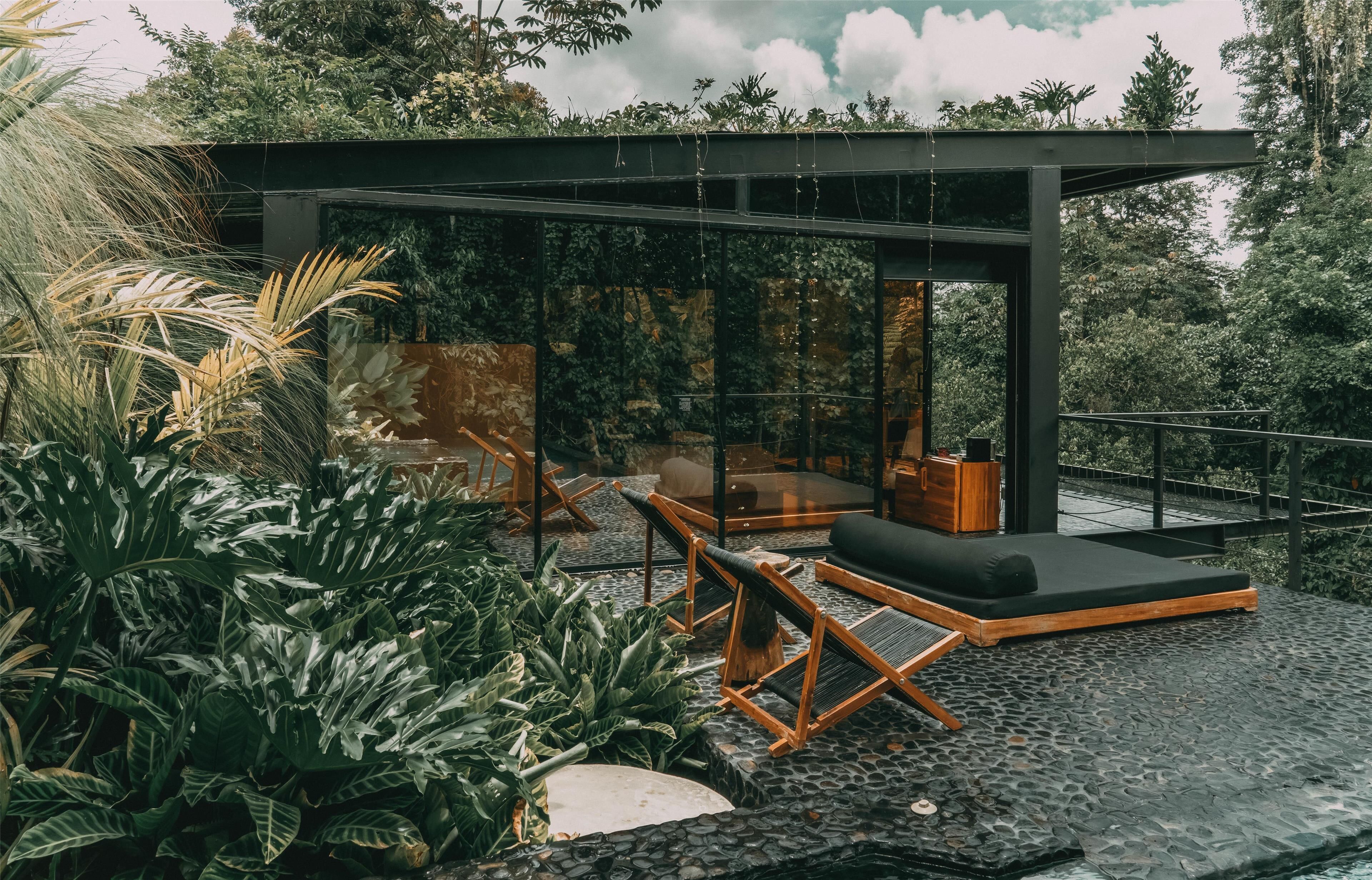 A modern outdoor lounge area with a glass enclosure, wooden furniture, and lush greenery.