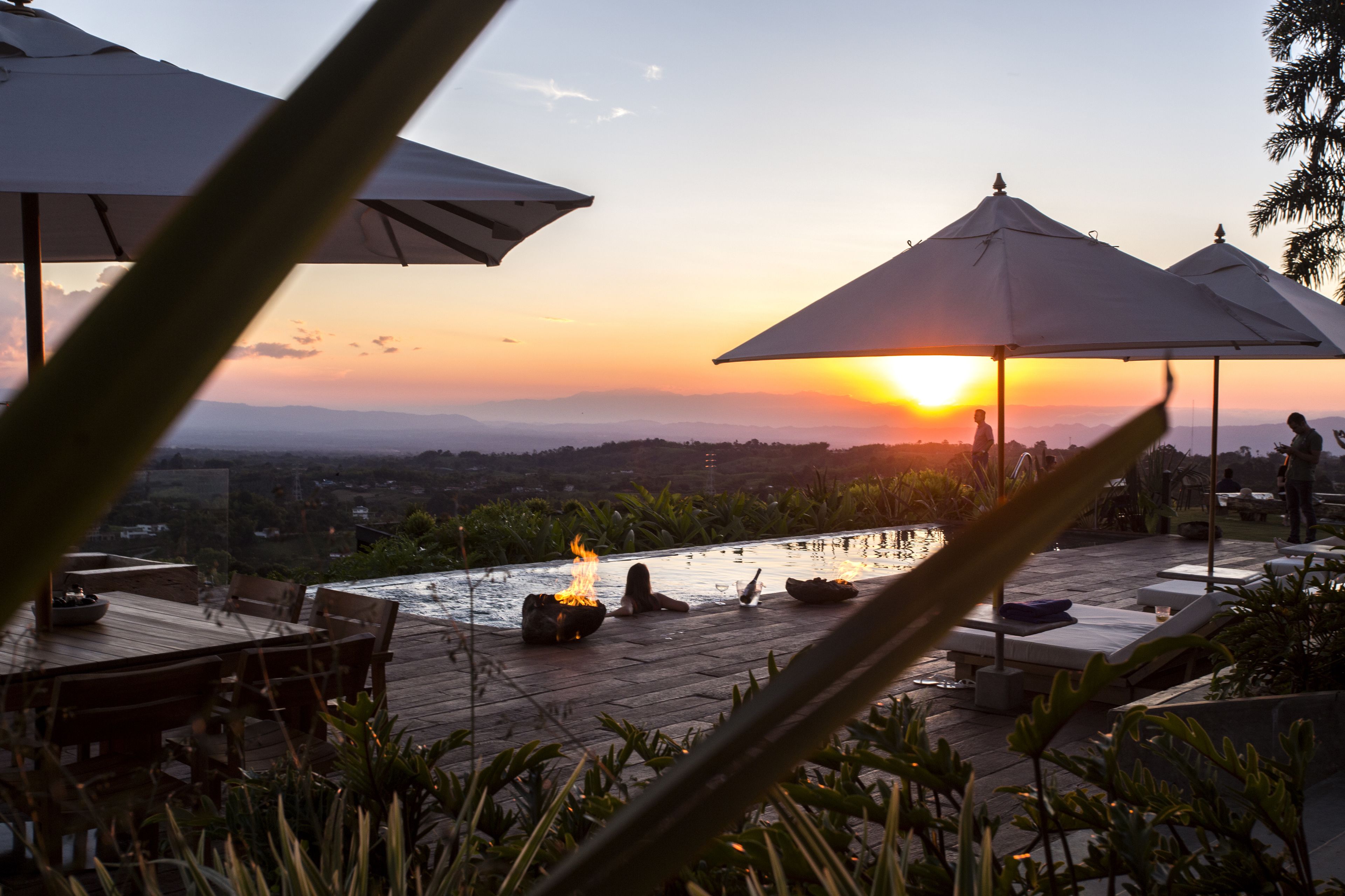 An outdoor pool area with lounge chairs and umbrellas, overlooking a scenic landscape during sunset.
