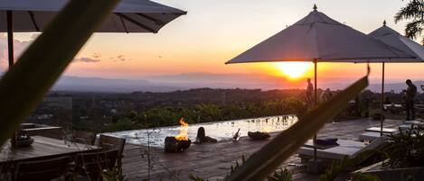 An outdoor pool area with lounge chairs and umbrellas, overlooking a scenic landscape during sunset.