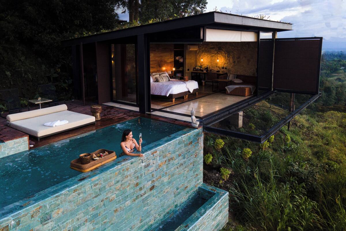 A luxurious outdoor infinity pool with a stone wall, a private bedroom with a bed and seating area, and a dining space with a table and chairs.