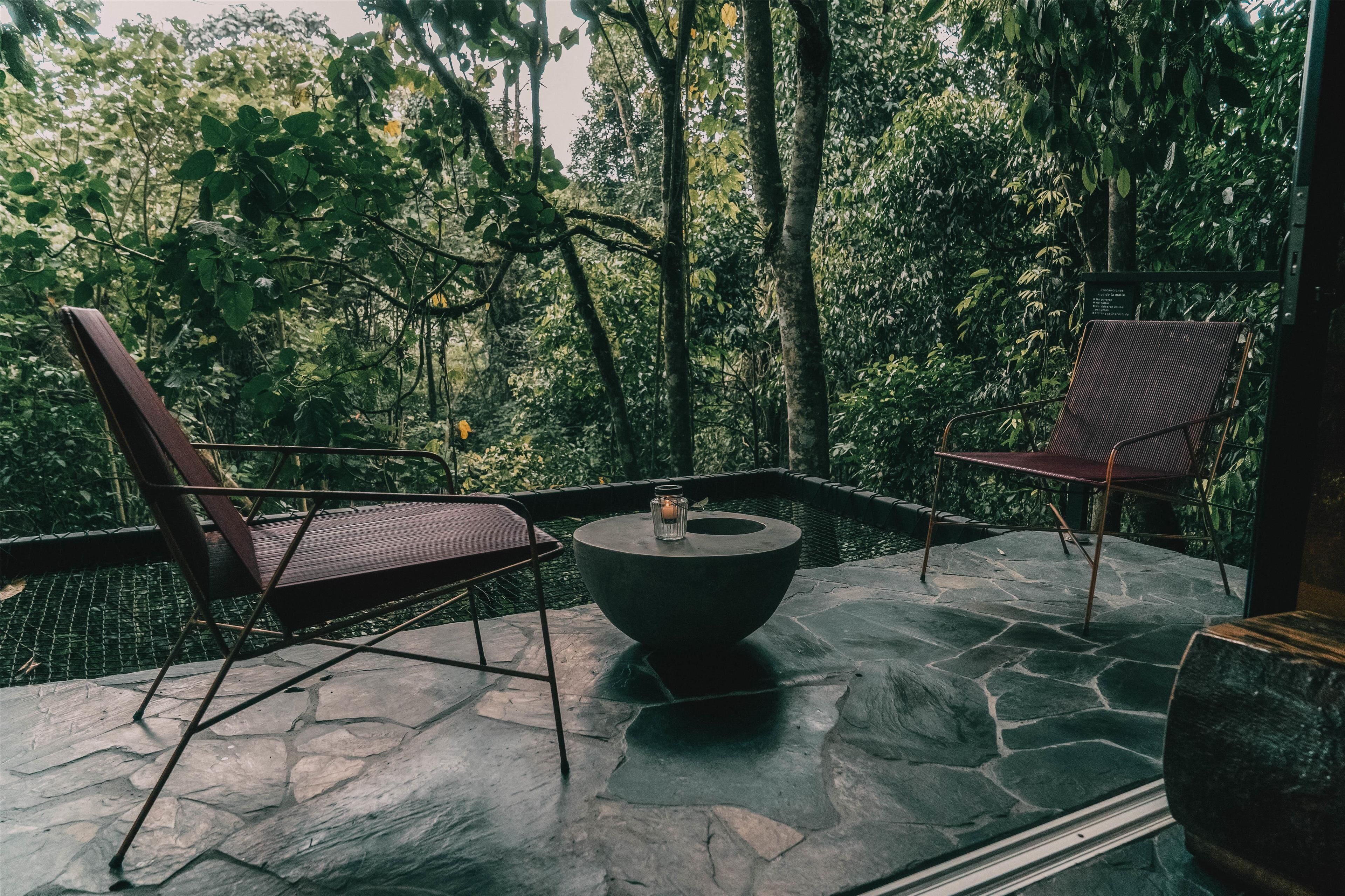 A stone-tiled patio with two wooden chairs and a round table, surrounded by lush greenery.