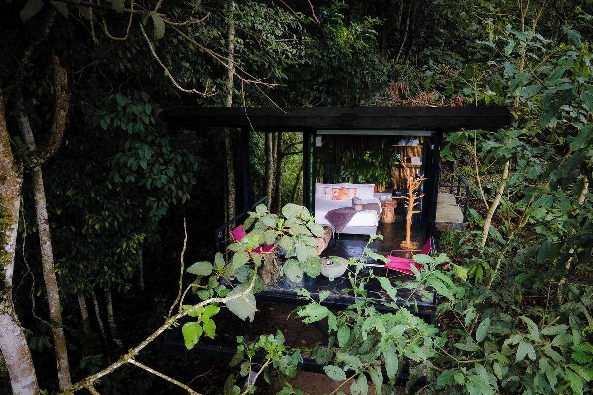 A secluded treehouse bedroom with a bed, a small table, and a chair, surrounded by dense foliage.