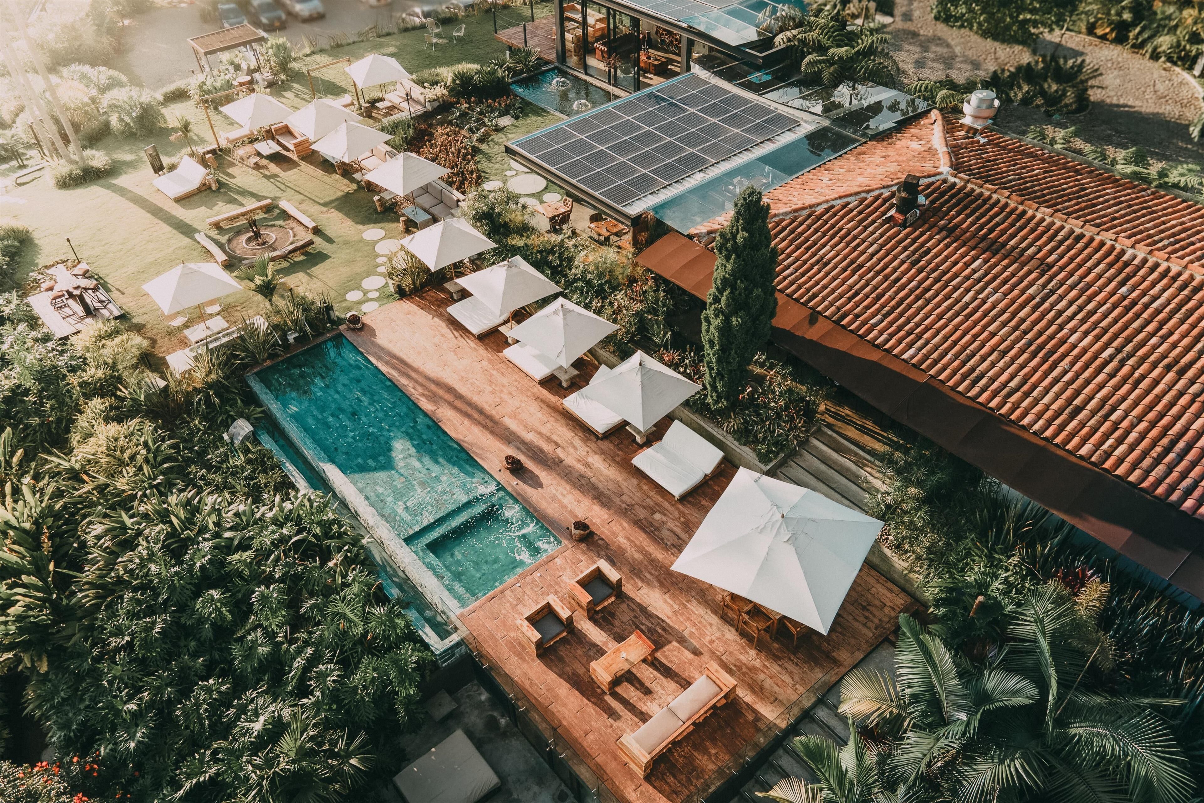 An aerial view of a resort with a swimming pool, sun loungers, umbrellas, and a solar panel.
