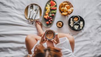 A person sitting on a bed with a plate of fruit, a bowl of cookies, and a cup of coffee.