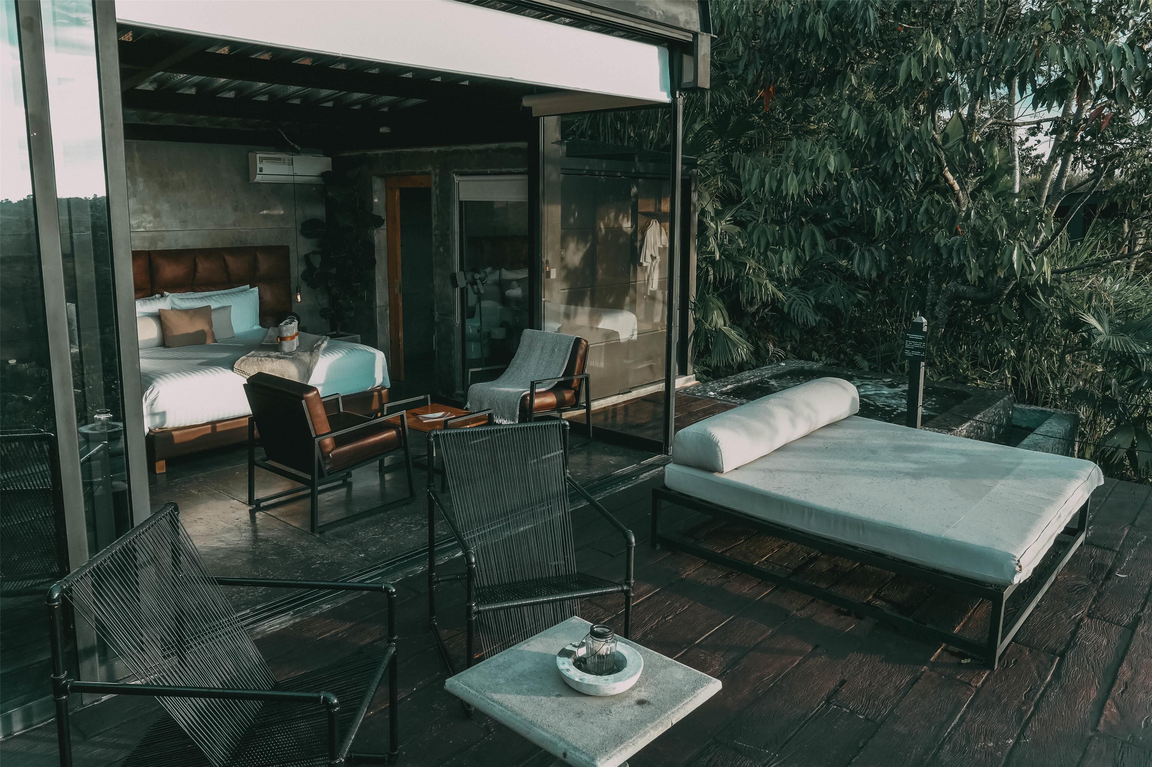 A modern outdoor bedroom with a bed, two chairs, a table, and a glass wall overlooking a lush garden.