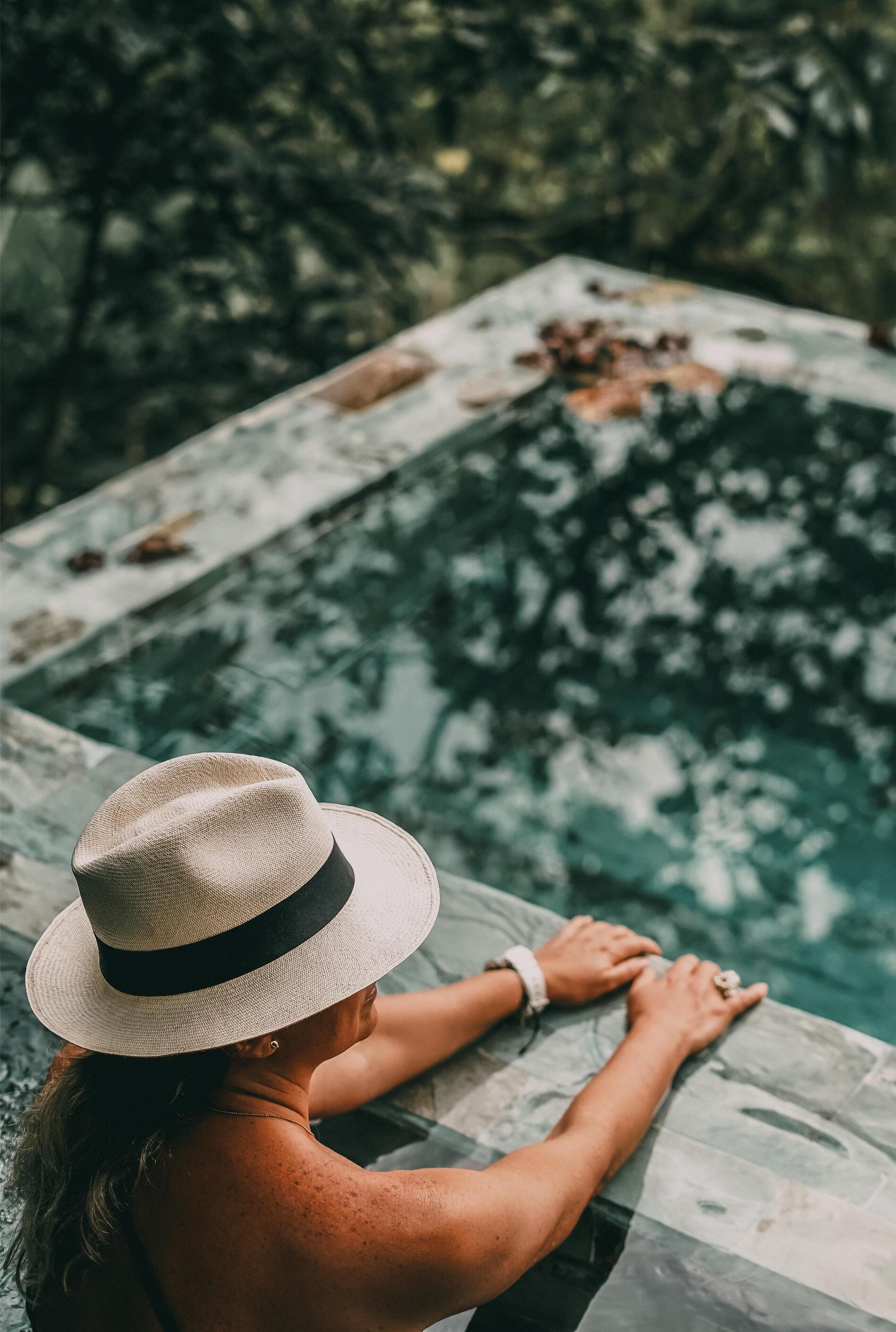 A person leaning on a pool edge with a hat, surrounded by a natural, outdoor setting.