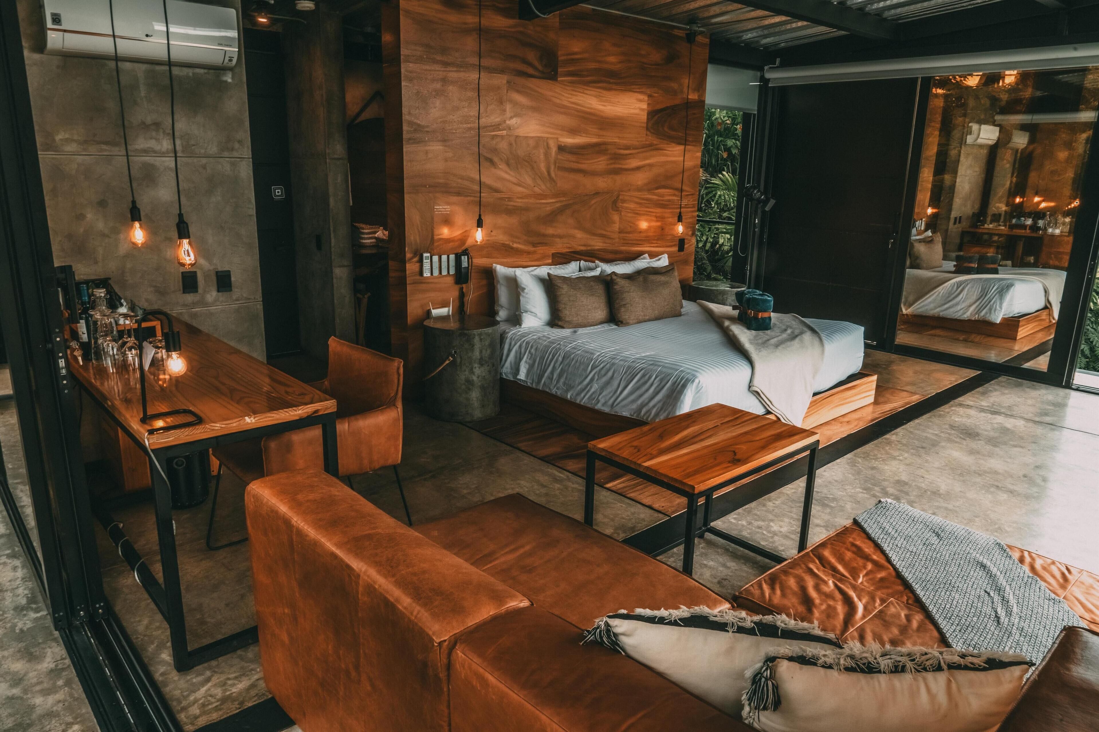 A modern, well-lit room with a wooden headboard bed, a wooden dining table, and a leather sofa.