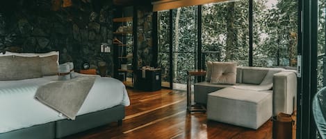 A spacious bedroom with a large bed, a sofa, and a coffee table. There is a stone wall and a large window offering a view of trees.