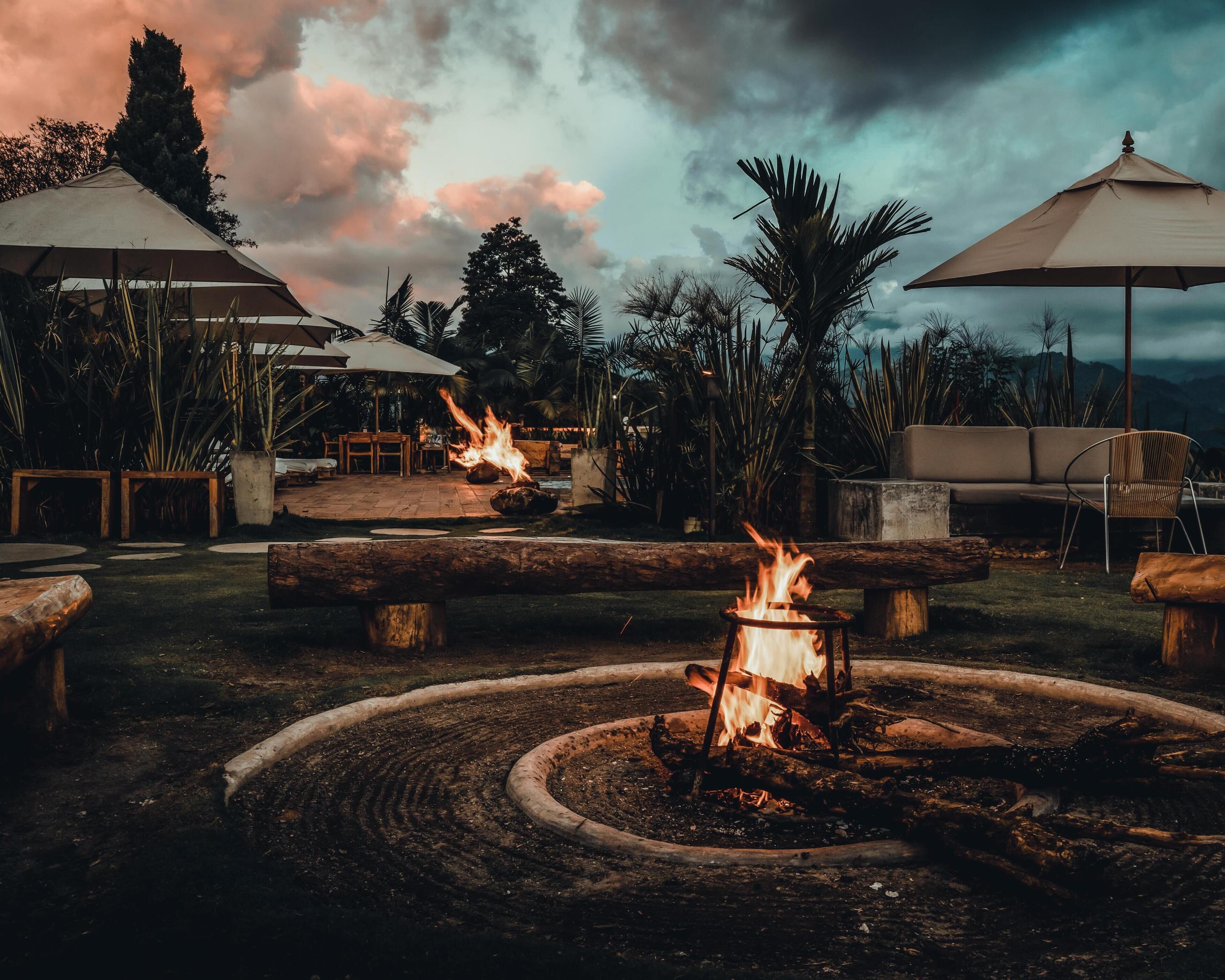 A fire pit with flames, surrounded by outdoor furniture and umbrellas, set against a backdrop of trees and a cloudy sky.