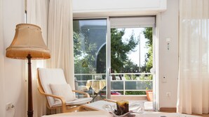 A hotel room with a large sliding glass door to a balcony, a wooden chair, a table with a lamp, and a bowl of fruit. - Fabulous Apartment Across Flisvos (Palaio Faliro)