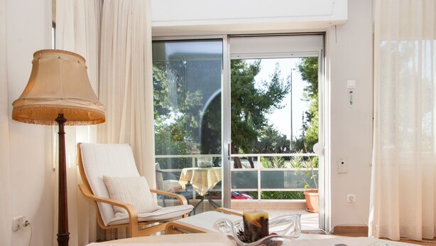 A hotel room with a large sliding glass door to a balcony, a wooden chair, a table with a lamp, and a bowl of fruit. - Fabulous Apartment Across Flisvos (Palaio Faliro)