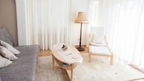 A modern room with a grey sofa, a small wooden table with a lamp, a white chair, and sheer curtains. - Fabulous Apartment Across Flisvos (Palaio Faliro)