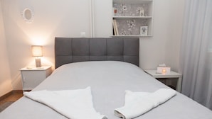 A neatly made bed with two folded towels, a bedside lamp, a small nightstand, and a shelf with decorative items. - Fabulous Apartment Across Flisvos (Palaio Faliro)