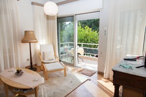 A room with a wooden table, a white chair, a lamp, and a view of greenery through a sliding glass door. - Fabulous Apartment Across Flisvos (Palaio Faliro)